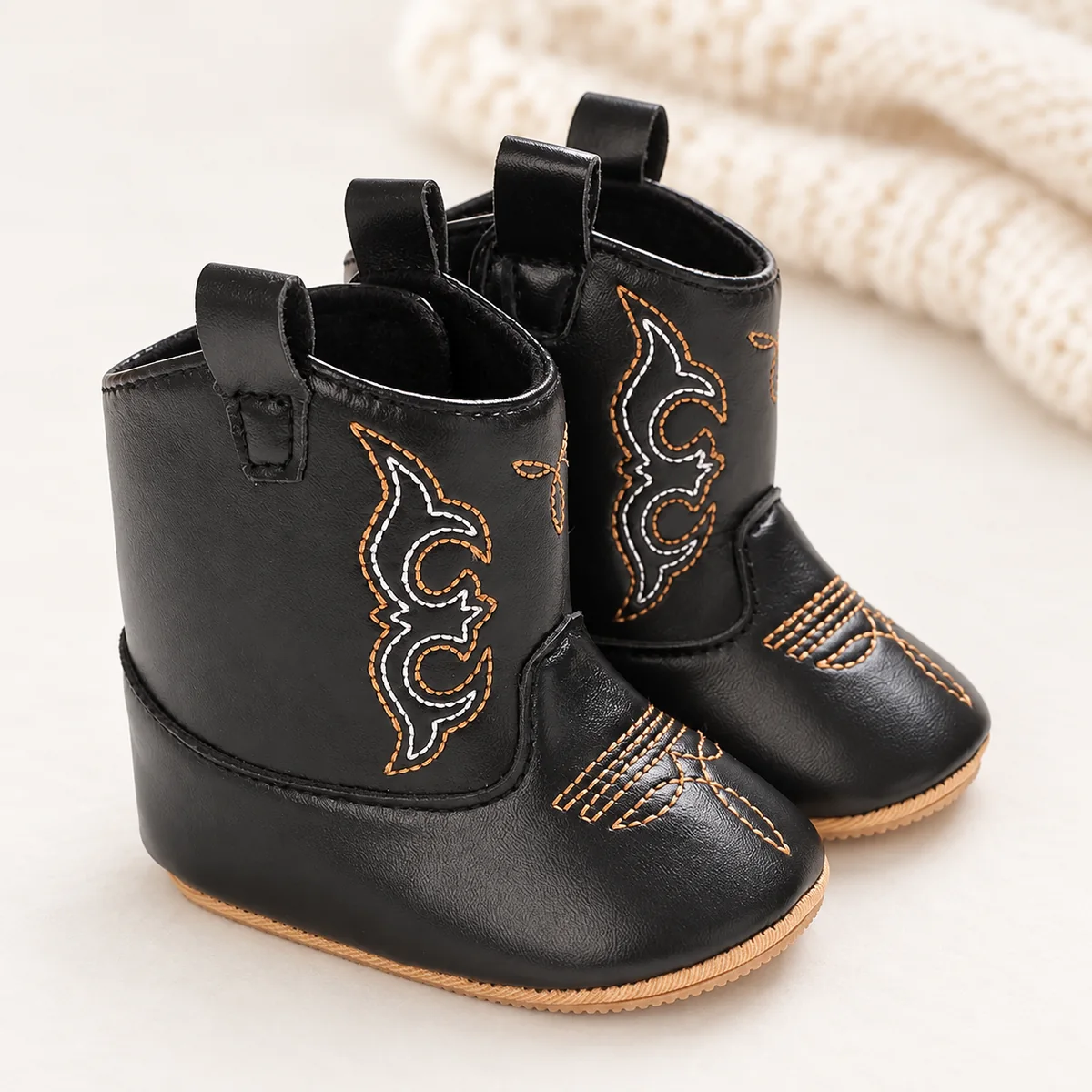 Bottines bébé western noires brodées arabesques sur un fond clair avec une maille écrue